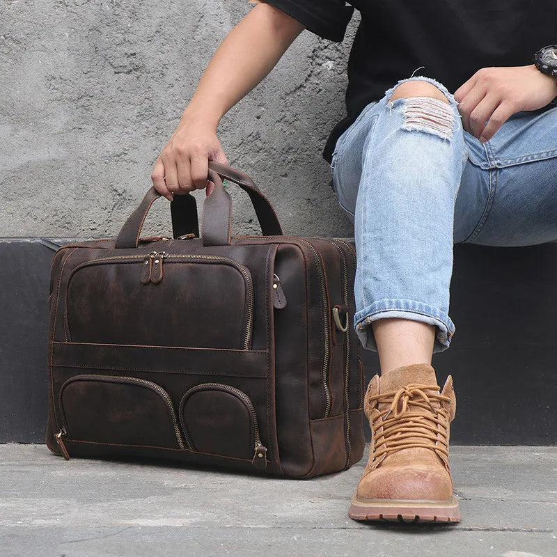 A person wearing a black shirt, ripped blue jeans, and tan hiking boots sits on a ledge against a grey wall. They are holding the handle of a large Leather 17" Business Travel Briefcase with multiple compartments and zippers placed beside them. The bag also features an adjustable strap for added convenience.