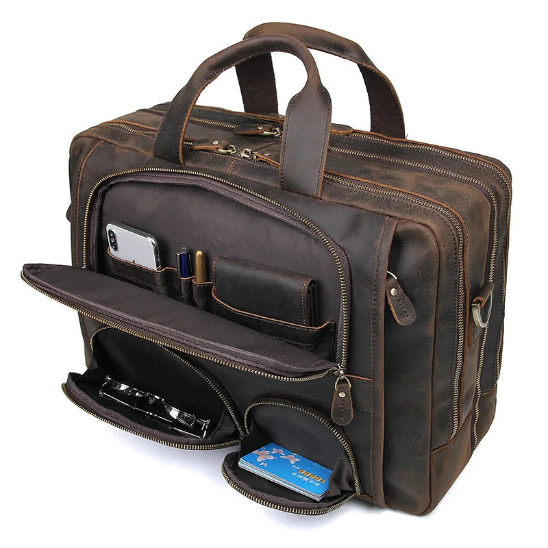 The Leather 17" Business Travel Briefcase is displayed in a rich brown genuine leather, featuring multiple compartments. The front pocket is open, showcasing an array of items such as a smartphone, pens, business cards, and a pair of sunglasses. The briefcase comes equipped with sturdy handles, durable zippers, an adjustable strap for convenience, and a dedicated laptop compartment.