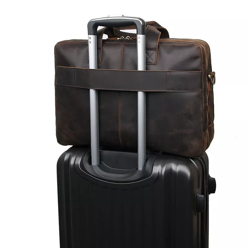 A black rolling suitcase with a telescoping handle has a Leather 17" Business Travel Briefcase securely strapped to it. The briefcase, featuring an adjustable strap and a dedicated laptop compartment, is attached by a rear strap slipped over the suitcase's handle, making it convenient for travel. The background is white.