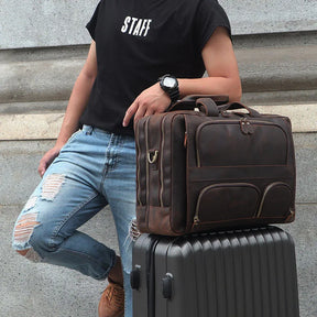 A person wearing a black "STAFF" t-shirt, distressed blue jeans, and a wristwatch leans against a stone wall. A brown genuine leather Leather 17" Business Travel Briefcase with an adjustable strap rests on top of a hard-shell suitcase, which the person has their hand on.