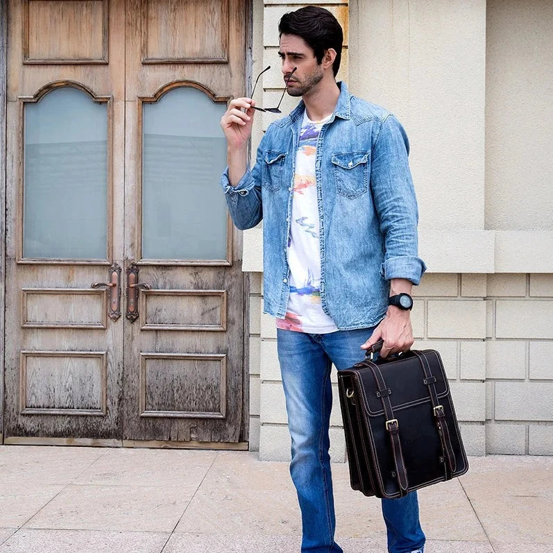 A man wearing a denim jacket over a tie-dye shirt and jeans stands in front of a wooden door, holding glasses in one hand and his Leather Business Travel Briefcase Backpack in the other. He appears to be deep in thought or waiting for someone.