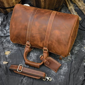 A Weekender 18 Leather Bag in brown, made from genuine cowhide leather, is showcased on a dark, textured fabric surface. The bag includes two short handles and a detachable shoulder strap with metal buckles. Its slightly distressed leather finish complements the spacious interior pockets, while leaves and wooden objects provide a backdrop.