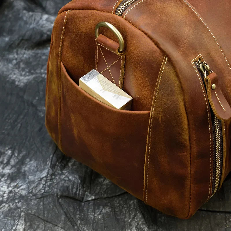 A Weekender 20 Leather Travel Bag made of genuine cowhide, featuring a gold zipper and a small front pocket holding a rectangular box, set on a dark, textured surface.