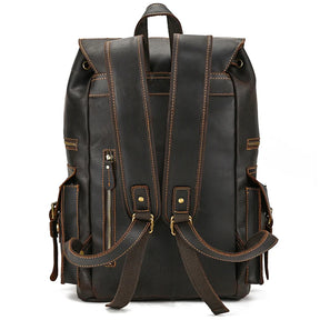 The Genuine Leather Travel Backpack, constructed from dark brown genuine cow leather, stands facing backward to highlight its two padded shoulder straps adorned with brass buckles and zippers. The design boasts multiple side pockets and detailed stitching throughout. A handle loop at the top ensures easy carrying, making it an ideal travel companion.