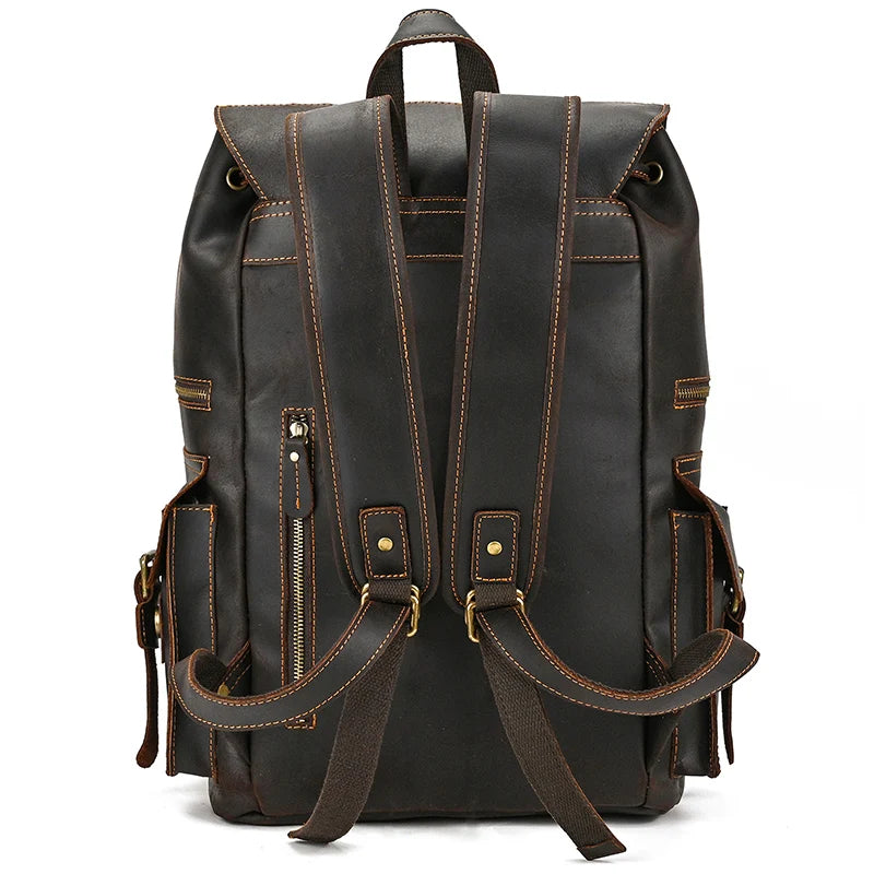 The Genuine Leather Travel Backpack, constructed from dark brown genuine cow leather, stands facing backward to highlight its two padded shoulder straps adorned with brass buckles and zippers. The design boasts multiple side pockets and detailed stitching throughout. A handle loop at the top ensures easy carrying, making it an ideal travel companion.