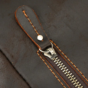 Close-up of a brown leather surface from the Genuine Leather Travel Backpack, showcasing an orange-stitched zipper. The zipper pull is crafted from matching brown leather with orange stitching and accented by a silver rivet. The metal zipper teeth and dark brown zipper tape are distinctly visible.