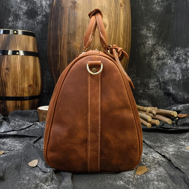 A brown Weekender 18 Leather Bag with a rounded, triangular shape stands upright against a rustic background. Made from genuine cowhide leather, the bag features a leather handle with a golden ring attachment. Behind it, wooden barrels and logs enhance the rugged, outdoorsy ambiance.