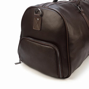 A close-up view of the Weekender Duffle 21 Leather Travel Bag reveals its dark brown genuine leather exterior with a side pocket. The travel bag features a textured surface, silver zippers, and a small metal ring detail on the side, all set against a white background.