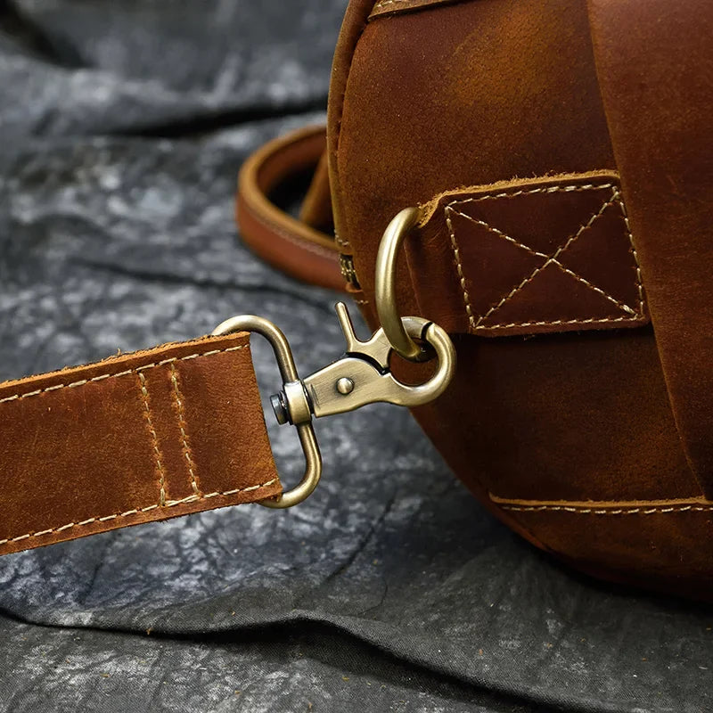 Close-up of the Weekender 20 Leather Travel Bag, crafted from genuine cowhide with intricate stitching. The image highlights a metal clasp and D-ring securing the adjustable strap to the bag's body. The backdrop showcases a dark, textured fabric.