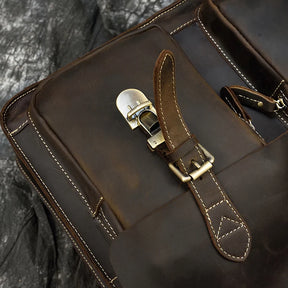 Close-up of the brown Leather 16" Business Briefcase against a dark, textured background. The focus is on the buckle and strap of the front pocket, highlighting the vintage, rugged design in genuine cowhide leather. White stitching is visible along the edges, adding contrast to the material.