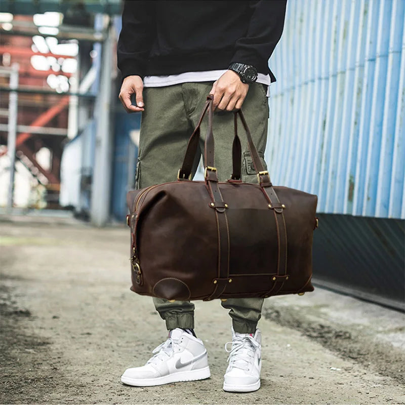 A person in a black long-sleeve shirt and green cargo pants is holding a large Weekender 20 Leather Travel Duffle made of genuine cowhide leather with both hands. The individual is also wearing white high-top sneakers and a wristwatch. The background features an industrial setting with a corrugated metal wall.