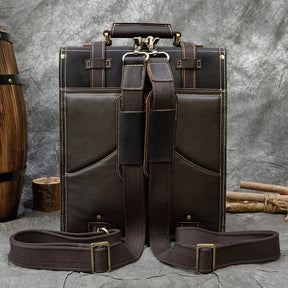 The Leather Business Travel Briefcase Backpack sits upright against a textured gray backdrop, exuding style and versatility. Crafted from genuine cowhide leather with light stitching, its dark brown hue stands out prominently. Two adjustable straps lay across the front. Nearby, wooden barrels and sticks add a rustic touch to the setting.