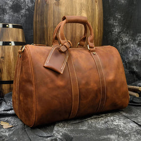 A large Weekender 18 Leather Bag, crafted from genuine cowhide leather, rests on a textured surface. The bag boasts sturdy handles, a luggage tag, and visible stitching. Featuring spacious interior pockets, it is placed among wooden barrels against a dark backdrop, evoking a rustic and vintage aesthetic.