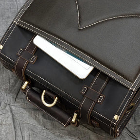 A close-up of the Leather Business Travel Briefcase Backpack with its flap partially open, revealing a white tablet inside. This versatile backpack features a dark exterior made of genuine cowhide leather, complemented by brown leather straps and brass buckles, all resting on a crinkled gray surface.