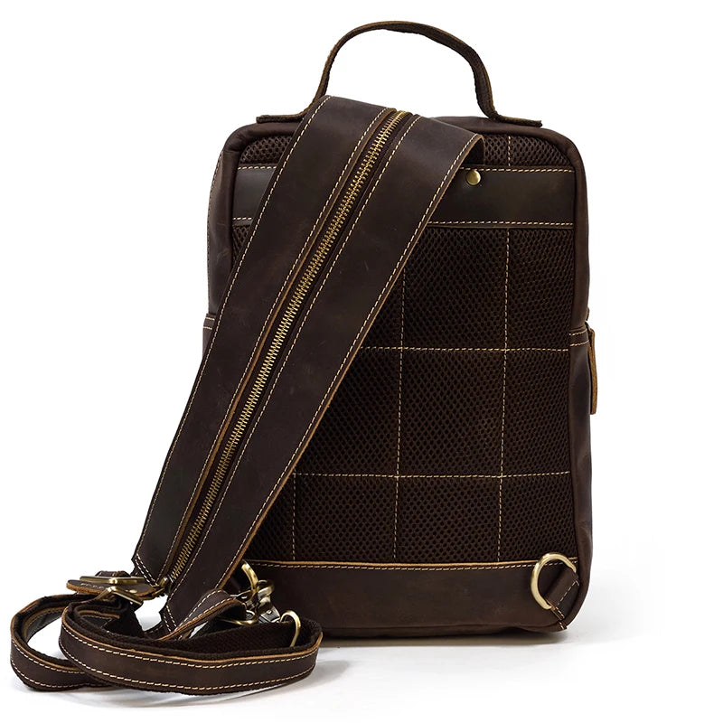 The Leather Travel Daypack Shoulder Bag is a dark brown, genuine cowhide leather backpack featuring brass hardware. This versatile daypack comes equipped with a top handle and a thick, adjustable shoulder strap along with multiple zippers. The front displays an elegant grid pattern with mesh detailing, and metal rings are positioned at the bottom corners for attachments.