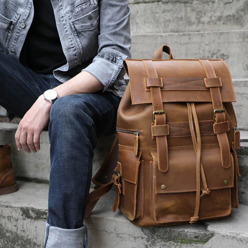 A person wearing jeans, a black shirt, and a gray jacket sits on concrete steps. Beside them is a Genuine Leather Travel Backpack made of durable cow leather with multiple pockets and buckle straps. The individual also sports a white-faced wristwatch that perfectly complements the vintage aesthetics of their ensemble.