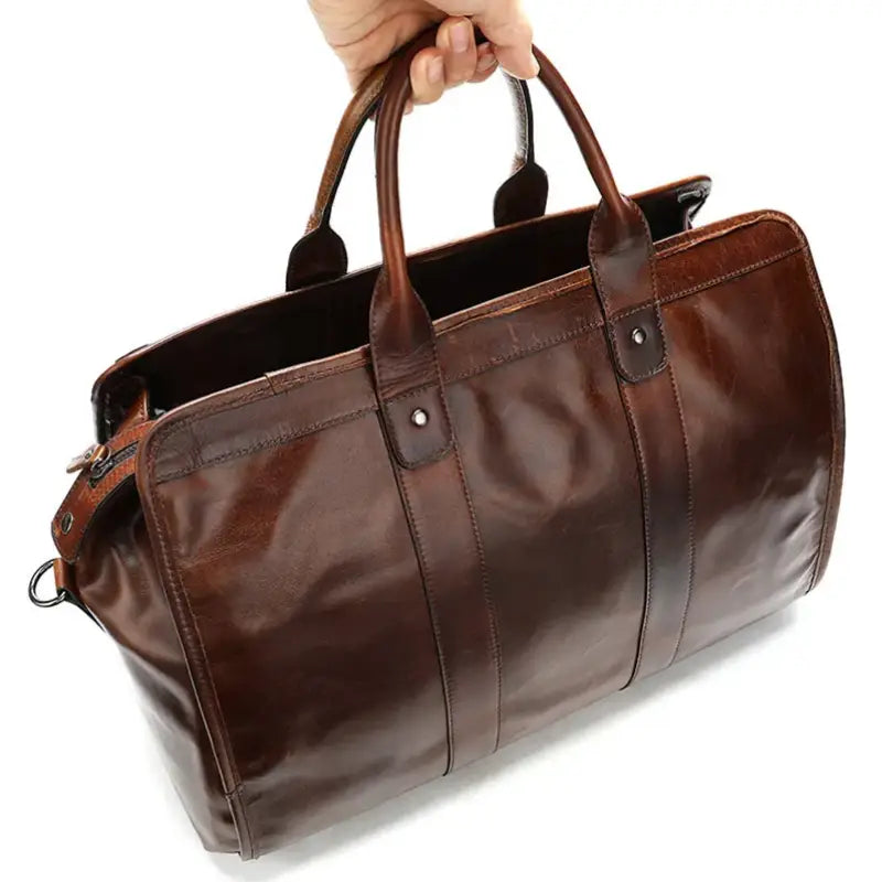 A person holding the Unisex Genuine Leather Travel Duffle Bag by its handles. The large, brown handcrafted bag features a structured design with visible stitching and metal hardware. The top is slightly open, revealing a glimpse of its spacious interior. The background is white.