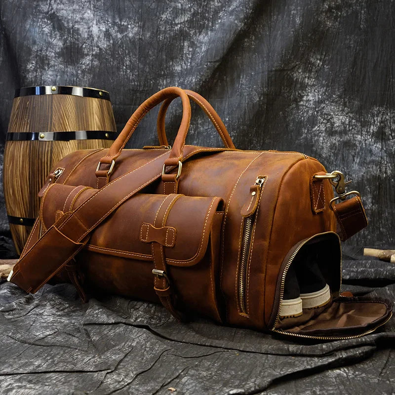 The Weekender 20 Leather Travel Bag, crafted from genuine cowhide leather and featuring multiple pockets, is displayed on a textured surface. This brown duffel bag includes a shoulder strap and a partially open shoe compartment that reveals a pair of white shoes. In the background, a wooden barrel can be seen.