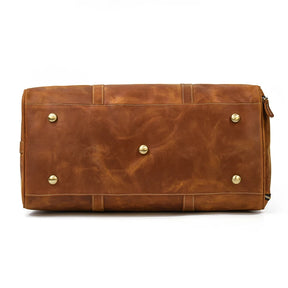 A brown Weekender 24 Duffle Bag, crafted from genuine cowhide leather, is shown from the bottom, featuring four metal studs for support. The bag has visible stitching and appears to be high-quality with a smooth finish. A zipper is partially visible on the right side—ideal for travelers.