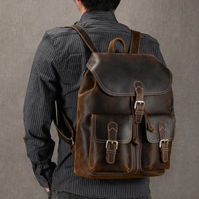 A person in a striped shirt carries a large, Genuine Leather Vintage Backpack featuring multiple pockets and buckle closures. The background is plain gray.