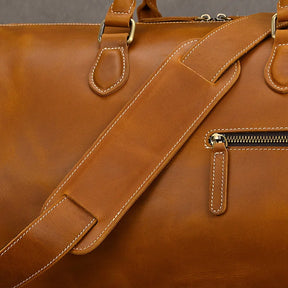 Close-up of the Leather Travel Duffle, a vintage brown leather bag with white stitching, featuring a strap and zippered pocket. The genuine leather has a smooth texture complemented by gold-toned hardware.