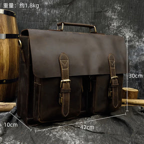 The Leather 16" Business Briefcase, crafted from genuine cowhide leather and featuring dual front buckles, is displayed against a rustic background. Measurements overlay the bag: width 42cm, height 30cm, depth 10cm, and a weight of approximately 1.8kg. An adjustable strap adds convenience while a wooden barrel and branches are partially visible beside it.