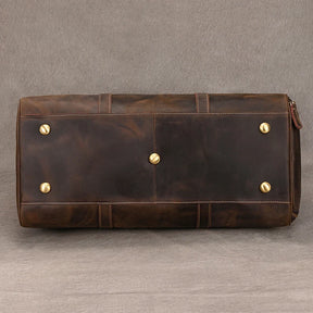 A close-up view of the bottom of a genuine cowhide **Weekender Duffle 22 Leather Travel Bag** with brass studs on a gray background. The leather has a slightly worn look, and the brass studs are placed evenly across the base for support and stability.