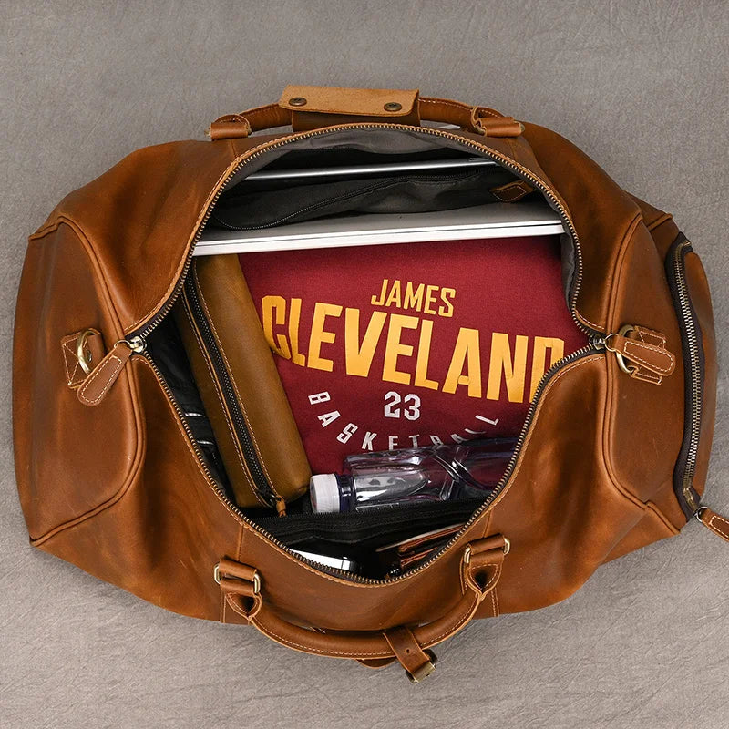 The Weekender Duffle 22 Leather Travel Bag is open, revealing its contents: a basketball jersey with "James" and "Cleveland 23" on it, a laptop, a brown leather wallet, a water bottle, a notebook, and a pair of sunglasses. The duffle bag is placed on a gray surface.