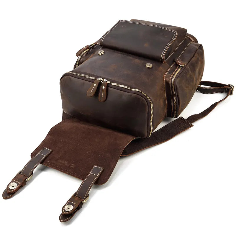 The Vintage Leather Travel Backpack in dark brown is made from genuine cowhide leather, featuring multiple zippered compartments and durable leather straps. The open front flap reveals a suede interior with a laptop compartment. It sits stylishly against a white background.