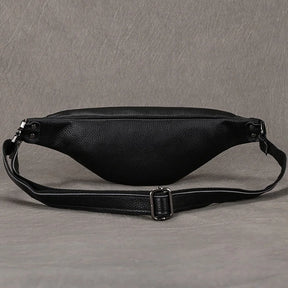 The Black Leather Belt Bag is displayed against a gray textured background. This black crossbody bag features a sleek, versatile design with a curved main compartment and an adjustable strap boasting a metal buckle. The premium leather material showcases a high-quality pebbled texture.