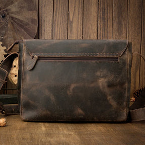 The **Vintage Leather Shoulder Bag** in brown rests on a wooden surface with a matching backdrop. Crafted from genuine cow leather, it boasts a front zippered pocket, a flap closure, and an adjustable strap with metal clasps. Its distressed, worn appearance enhances its timeless style.