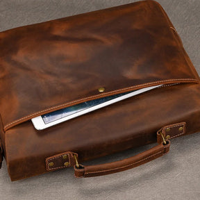 The Leather 14" Briefcase Shoulder Bag in brown leather is partially open, revealing a tablet inside. Crafted from genuine cowhide, the briefcase exudes professional style with its handle and brass hardware. It rests elegantly on a grey surface.