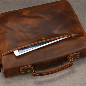 The Leather 14" Briefcase Shoulder Bag, made from genuine brown cowhide leather, is partially open to reveal a tablet inside. The briefcase features a handle and a visible snap button closure, exuding professional style. The polished leather, with its slightly worn look, enhances its character. It is placed on a gray surface.