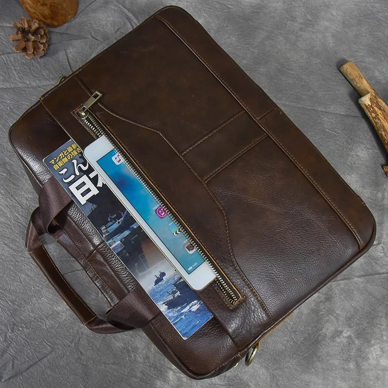 A 16" Brown Leather Briefcase is placed on a grey surface. The briefcase's front pocket is unzipped, revealing a partially visible Japanese magazine and a smartphone. A pine cone and a piece of wood are placed nearby, along with the adjustable strap neatly coiled beside it.