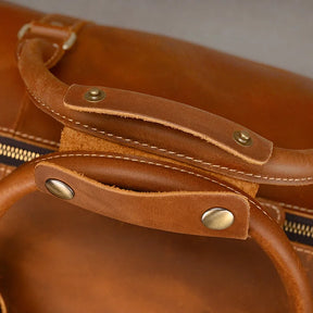 Close-up of the handles and zipper of a brown Weekender Duffle 22 Leather Travel Bag. The handles are secured with brass hardware, and the texture of the leather is visible. The image focuses on the craftsmanship and detailing of this premium leather travel bag.