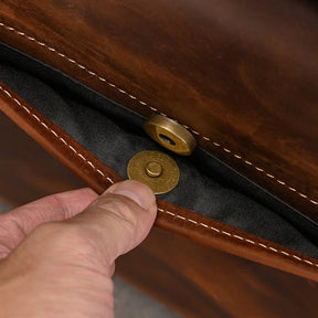 A close-up image showcases a person's hand opening the Leather 14" Briefcase Shoulder Bag, crafted from genuine cowhide leather with contrasting white stitching. The bag's black interior lining and brass magnetic snap closure, with both parts of the fastening mechanism visible, epitomize professional style.