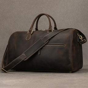 The Leather Travel Duffle, a vintage-inspired dark brown genuine leather bag, features sturdy handles, a detachable shoulder strap, a front zip pocket, and brass hardware. It is set against a simple gray background, blending ruggedness with sophistication.