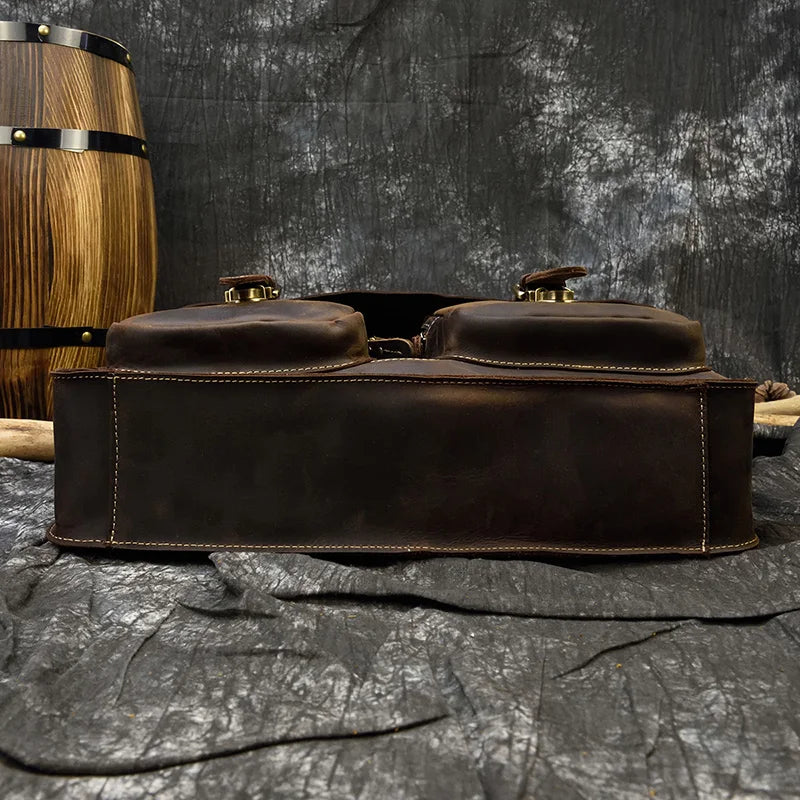 The 16" Leather Business Briefcase, featuring adjustable straps and dual compartments secured by buckles, lies flat on a dark, textured surface. In the background, a partially visible wooden barrel complements the scene, as the mottled grey and black backdrop enhances the rustic ambiance.