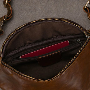 A brown Leather Belt Bag with a zipper opening is shown. Crafted from genuine cowhide leather, it features durable alloy hardware for added strength. Inside, there is a small zippered pocket containing a maroon item, possibly a passport or notebook. The bag's interior lining is dark brown; the exterior is smooth and slightly glossy.