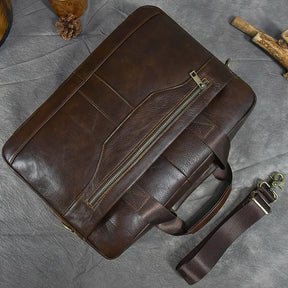 The 16" Brown Leather Briefcase, made from genuine dark brown leather, rests on a gray surface. It features a front zipper pocket, two top handles, and an adjustable shoulder strap with brass hardware placed next to the bag. Pinecones and pieces of wood are arranged nearby for decoration.