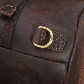 Close-up of the Vintage Leather Travel Backpack, crafted from genuine brown cowhide and featuring a brass D-ring with reinforced stitching. The leather has a slightly worn texture, and you can see a dark fabric strap above the D-ring, making it perfect for the stylish traveler.