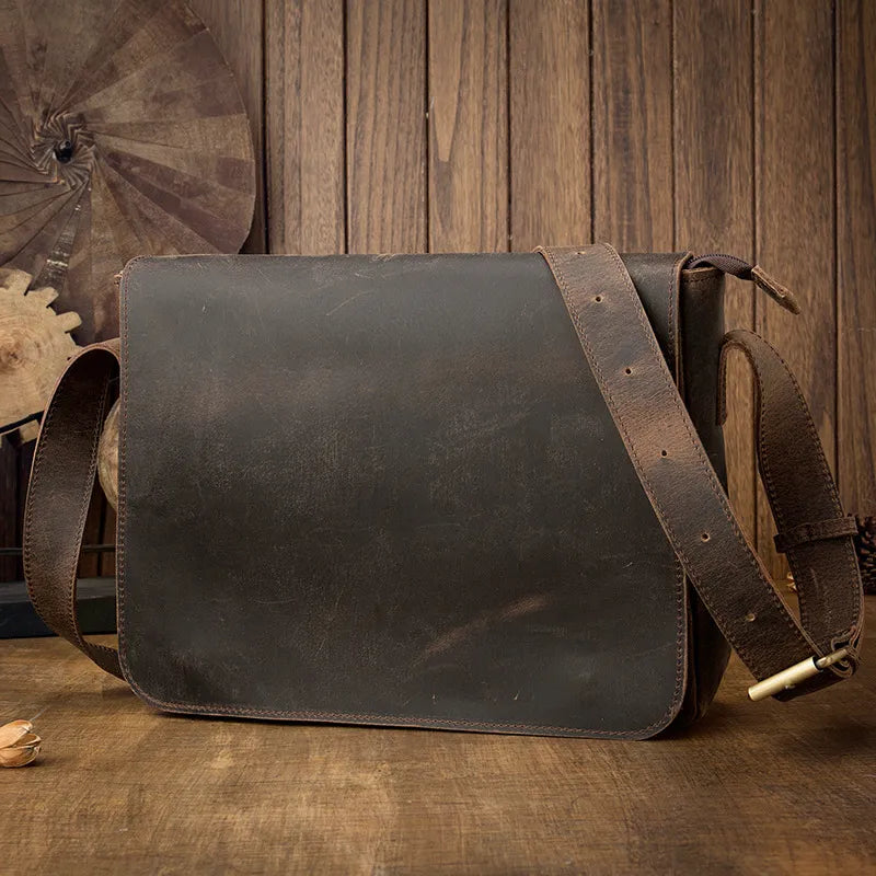 A Vintage Leather Shoulder Bag with a matching adjustable strap is displayed on a wooden surface against a backdrop of vertical wooden planks. Crafted from genuine cow leather, the bag exudes timeless style and a rustic, vintage appearance.