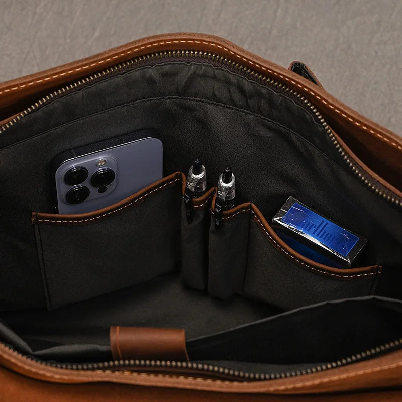 Interior view of the "Leather 14" Briefcase Shoulder Bag" made from genuine brown cowhide leather with a black lining, showcasing organized compartments holding a smartphone, two pens, and a blue packet of tissues. Its professional style is perfect for any business setting.