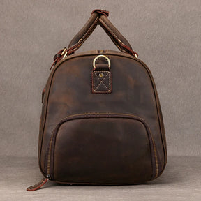 The Weekender Duffle 22 Leather Travel Bag stands upright against a neutral background. It features a zippered front pocket, a top handle with leather wraps, and gold-colored hardware, including a ring detail on the front. The genuine cowhide leather has a slightly worn, rustic finish.