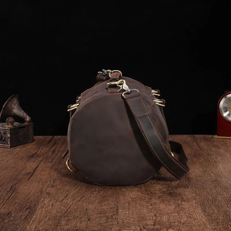 The Vintage Leather Travel Backpack, made from genuine cowhide leather with brass fixtures, is ideal for the fashionable traveler. It boasts an adjustable strap and is elegantly placed on a rustic wooden surface against a dark background, accompanied by a classic decor piece and clock partially visible.