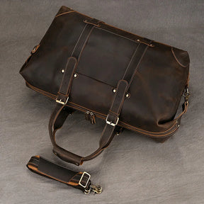 The Weekender 20 Leather Travel Duffle, a large, dark brown leather bag with a removable strap, rests on a gray surface. Featuring sturdy handles, silver hardware, and visible stitching, it exudes a rugged yet sophisticated appearance. Crafted from genuine cowhide leather, this duffle promises durability and style for any journey.