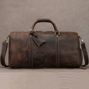 The Weekender Duffle 22 Leather Travel Bag is a rustic brown, genuine cowhide leather duffel bag featuring sturdy handles and a detachable shoulder strap. It showcases two parallel dark straps on the front with a small attached leather tag, resting gracefully against a neutral background.