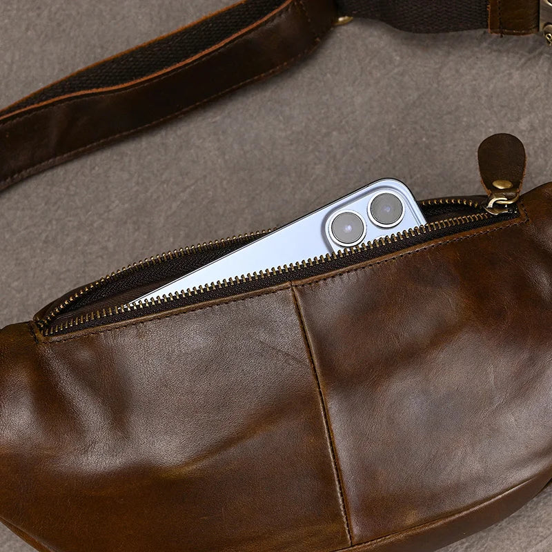 A brown Leather Belt Bag with a metal zipper is partially open, revealing a silver smartphone with dual rear cameras inside. Crafted from genuine cowhide leather, the bag is laid flat on a grey surface.