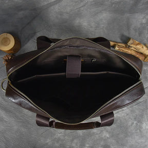 Top-down view of the open 16" Brown Leather Briefcase with a zippered main compartment. The interior features multiple sections, a padded laptop sleeve secured with a Velcro strap, and a zippered pocket. An adjustable strap is attached. The background includes a piece of wood, a pine cone, and a textured surface.