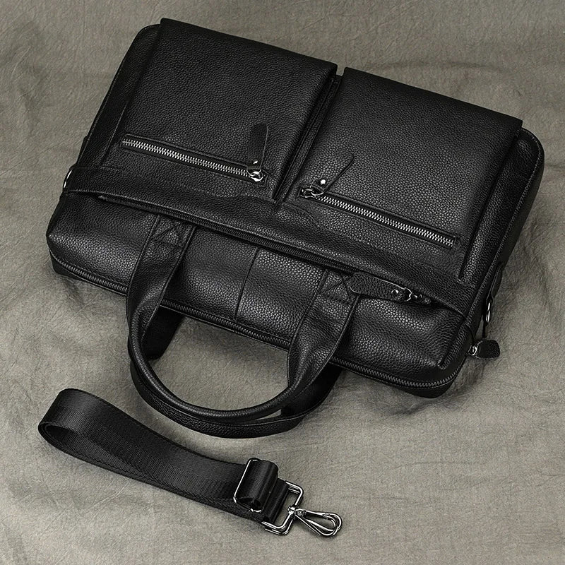 The 15" Black Briefcase, crafted from genuine black leather, features two front zippered pockets, dual handles, and an adjustable shoulder strap with a metal clasp, resting on a neutral-colored surface.